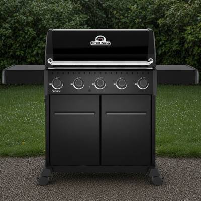 Black Broil King Crown 520 Shadow gas grill on gravel, lid closed, five knobs above two cabinet doors, hedge behind